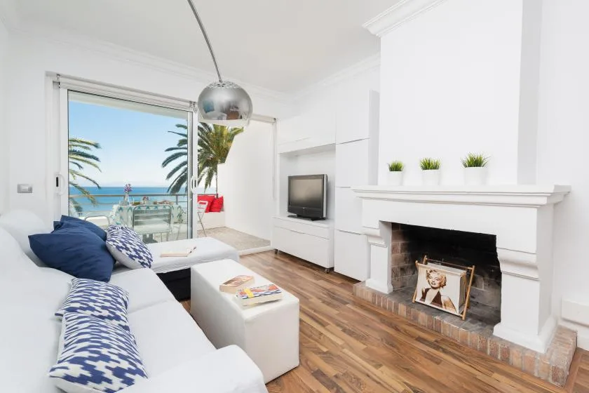 Una sala luminosa tiene asientos blancos, una chimenea y vistas al mar a través de puertas corredizas.