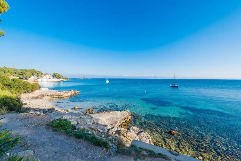 Una costa rocosa bordea aguas azules y claras, con barcos a lo lejos.