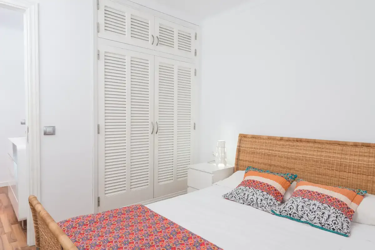 Un dormitorio luminoso con una cama, cojines estampados y puertas blancas de armario con persianas.