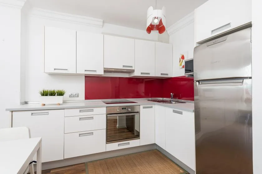 Una cocina blanca y luminosa con salpicadero rojo, encimeras y electrodomésticos de acero inoxidable.
