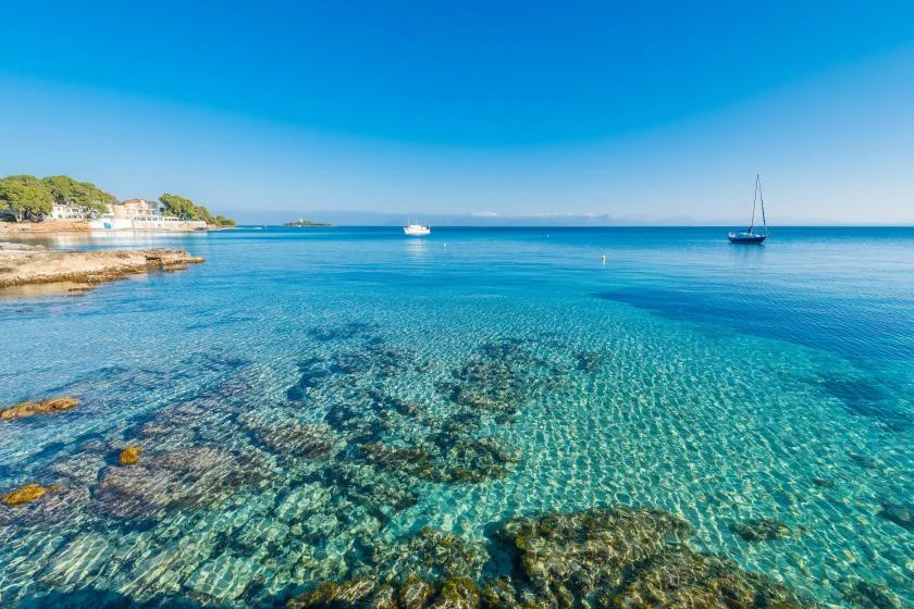 Una bahía azul y transparente con aguas poco profundas y rocosas, costa y barcos.