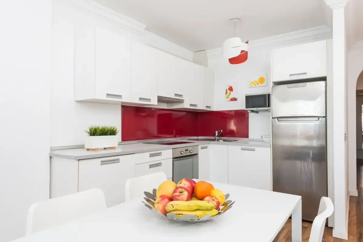 Una cocina luminosa con gabinetes blancos, salpicadero rojo y una mesa con fruta.