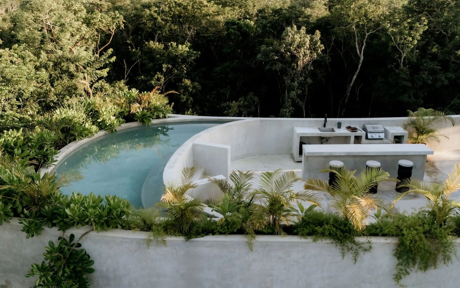 Una piscina al aire libre se curva junto a una barra con taburetes, rodeada de vegetación.