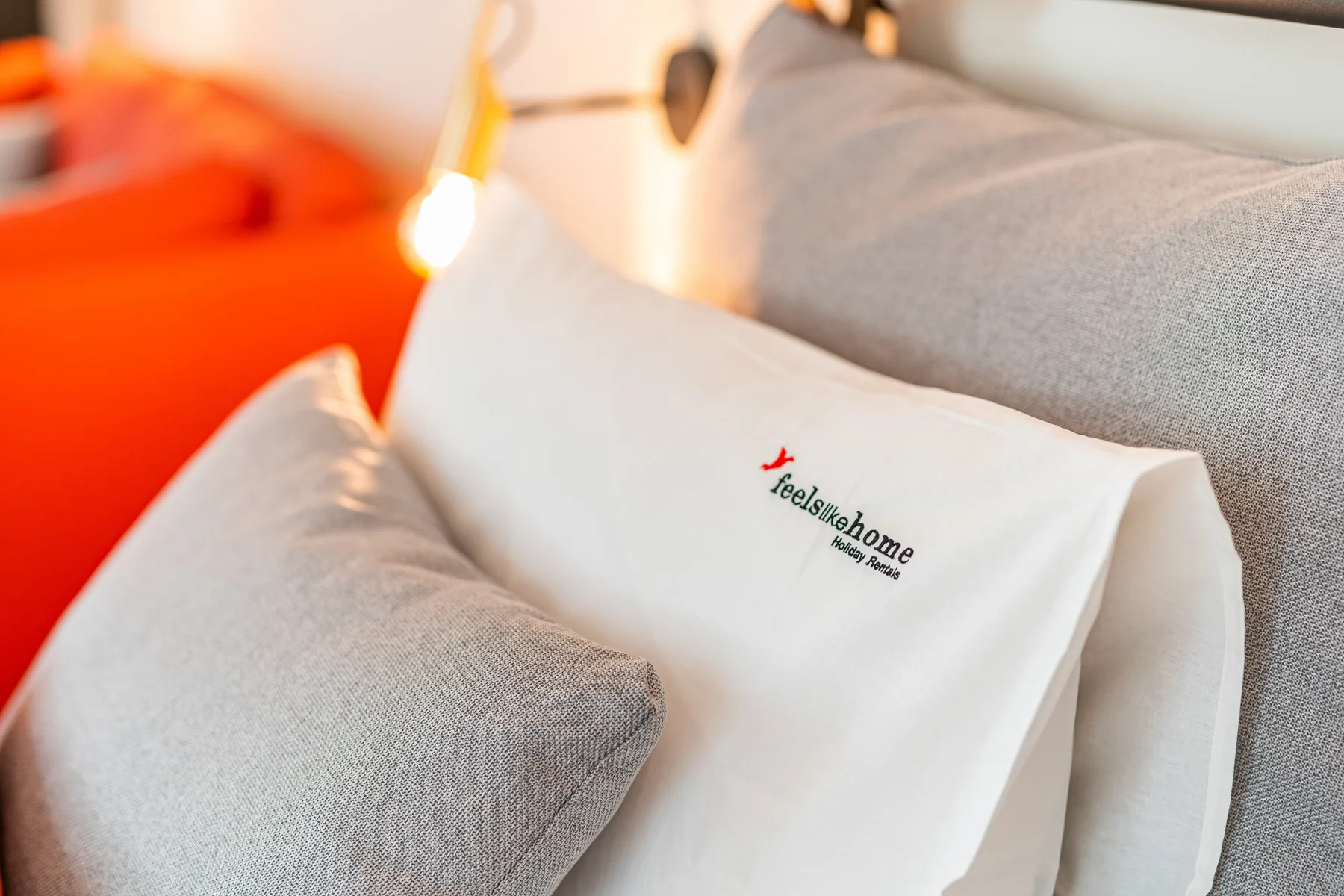 Una cama con ropa de cama blanca y almohadas grises está junto a una superficie naranja brillante.