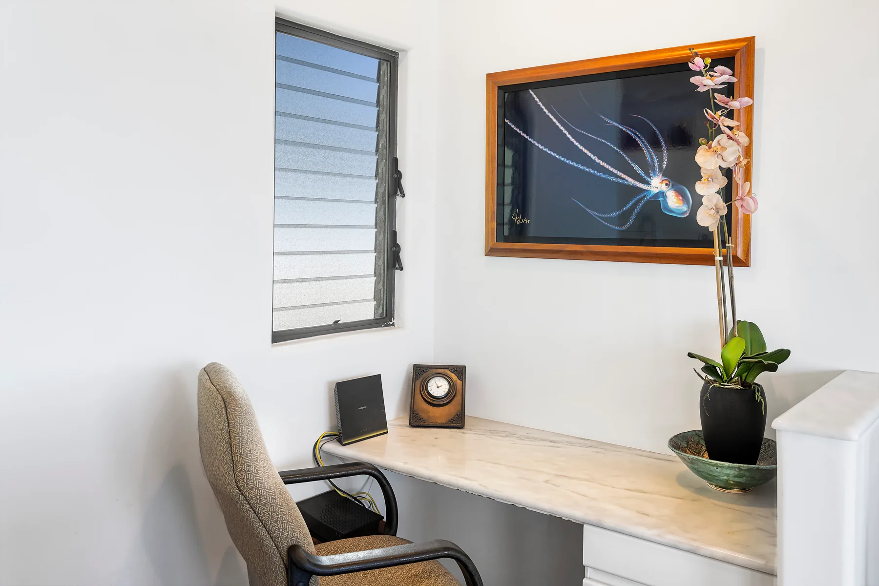 Un espacio de escritorio luminoso con una ventana, una obra enmarcada y una planta con flores.