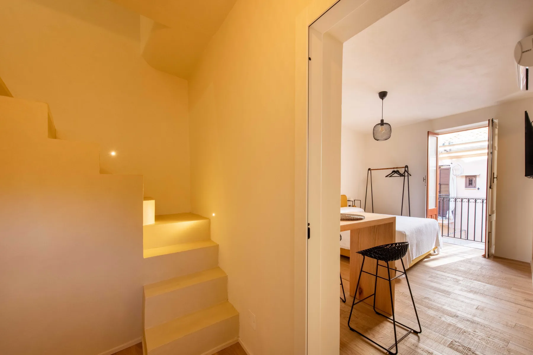 Una escalera de tonos cálidos da a un dormitorio con escritorio y puerta al balcón.