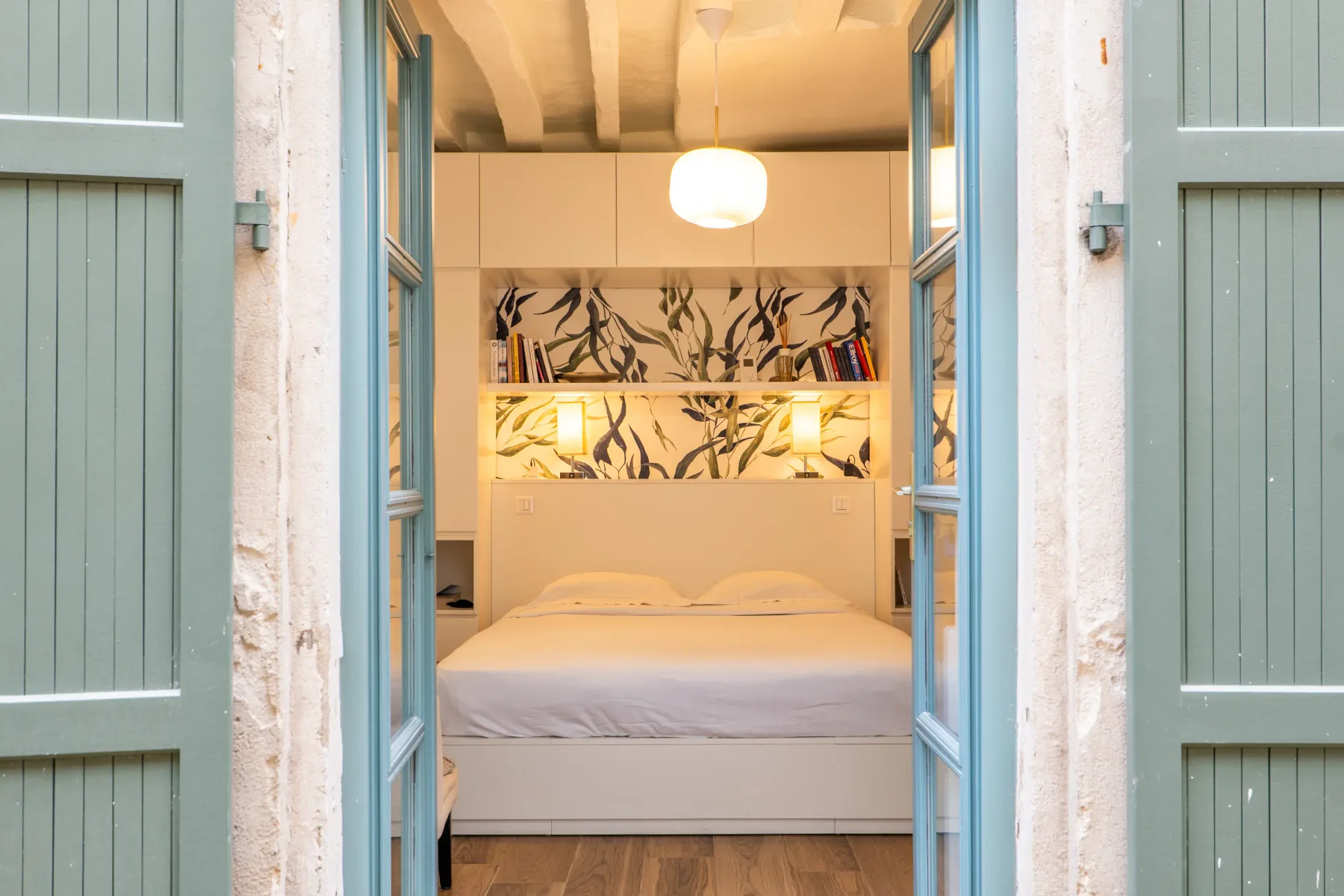 A bedroom with a bed, built-in shelves, and open blue shutters.