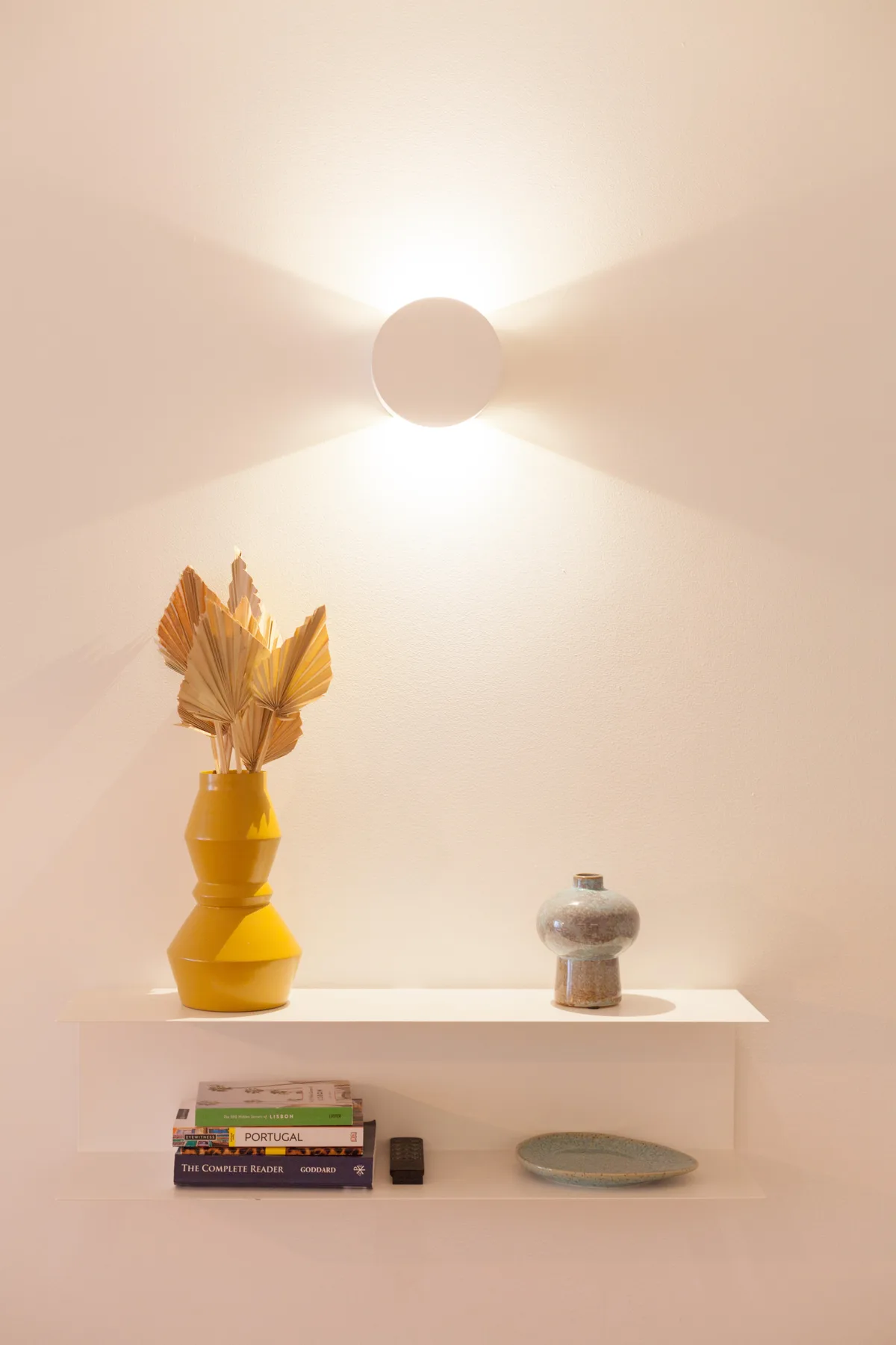 Un estante de pared muestra libros, un jarrón y un plato decorativo bajo una luz encendida.