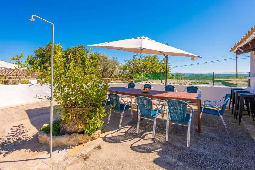 A sunny outdoor dining area has a long table beneath a large umbrella.