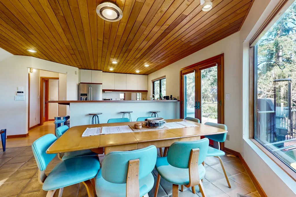 A dining area with turquoise seating and a long table beside large windows.