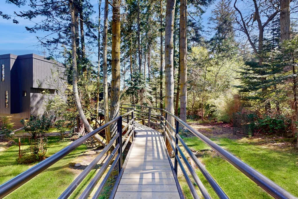 A raised walkway with railings crosses a wooded garden beside a dark building.