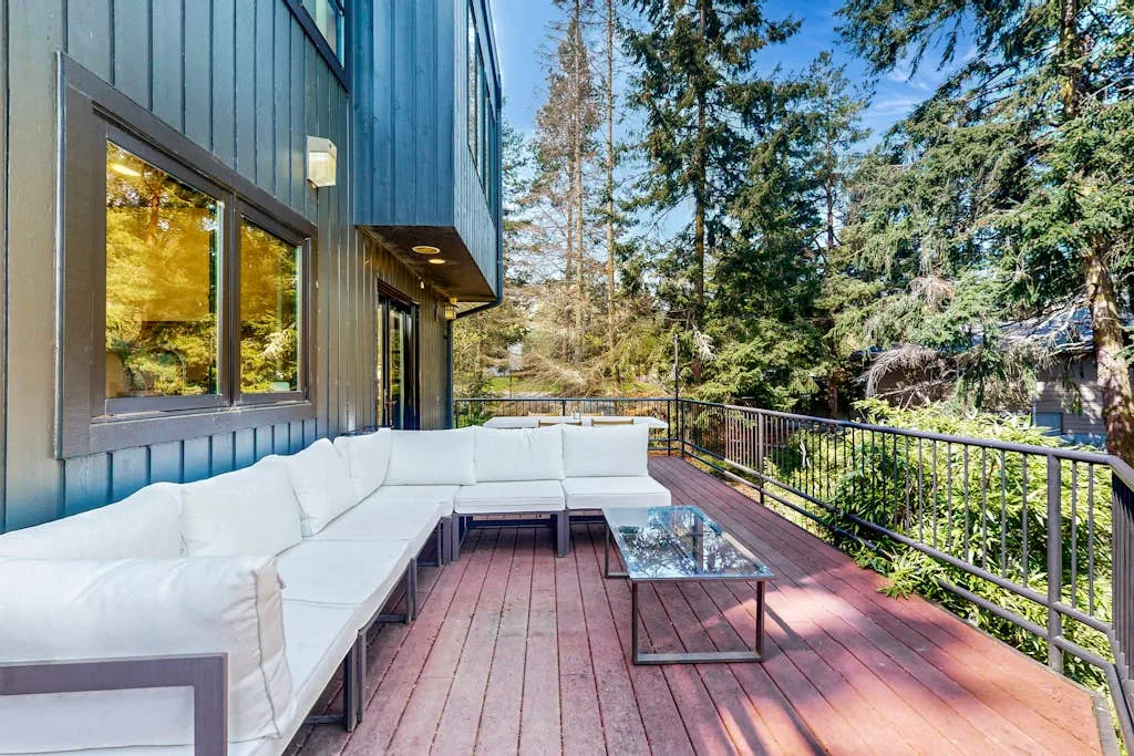 An outdoor deck has white seating and a glass-topped table beside a wooded view.