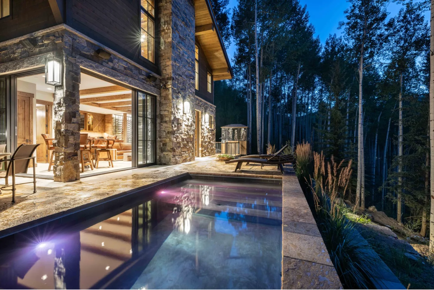 A modern house opens onto a lit outdoor pool beside a wooded area at dusk.