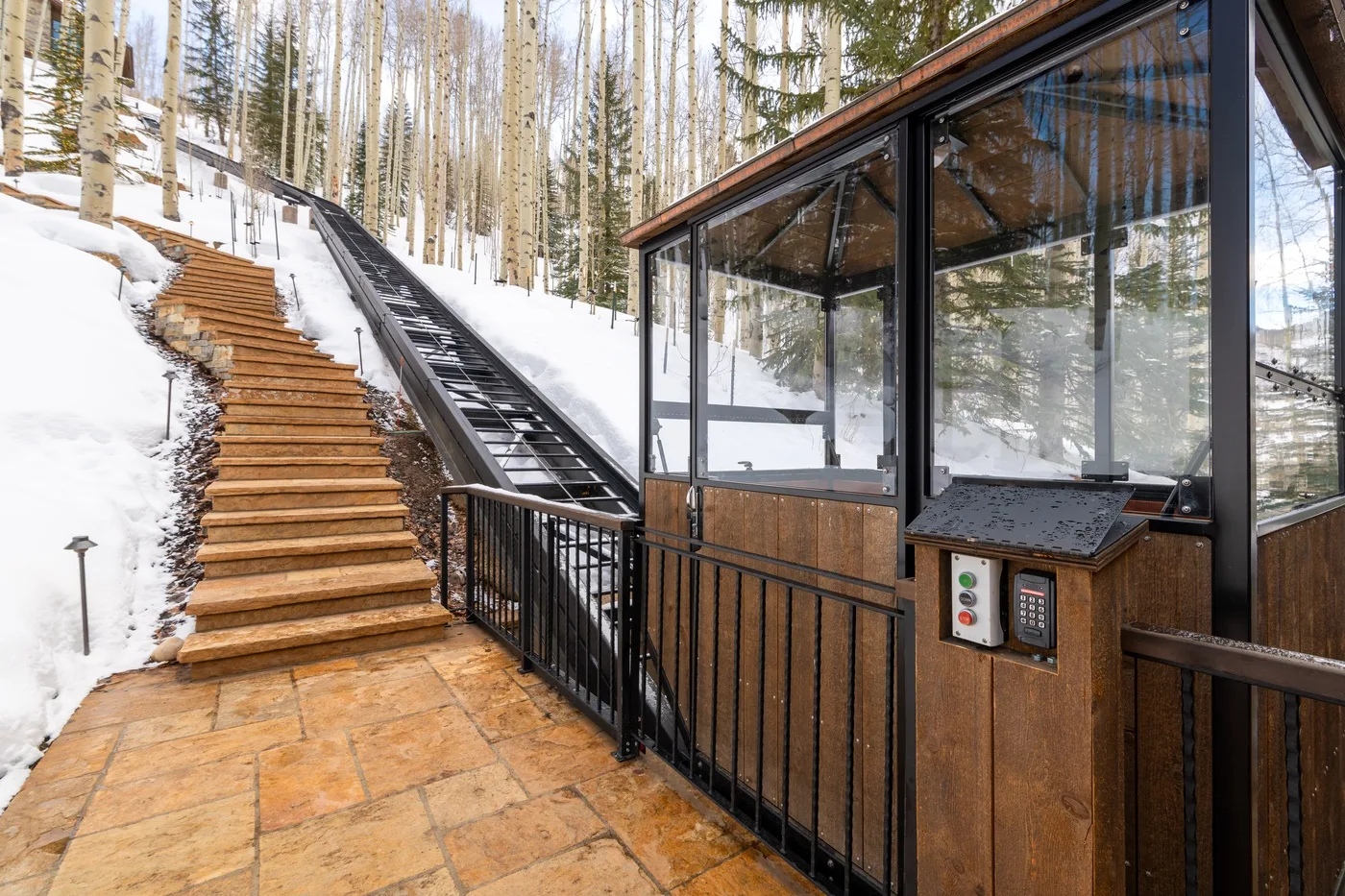 A glass-enclosed lift stands beside snow-covered outdoor stairs in a wooded area.