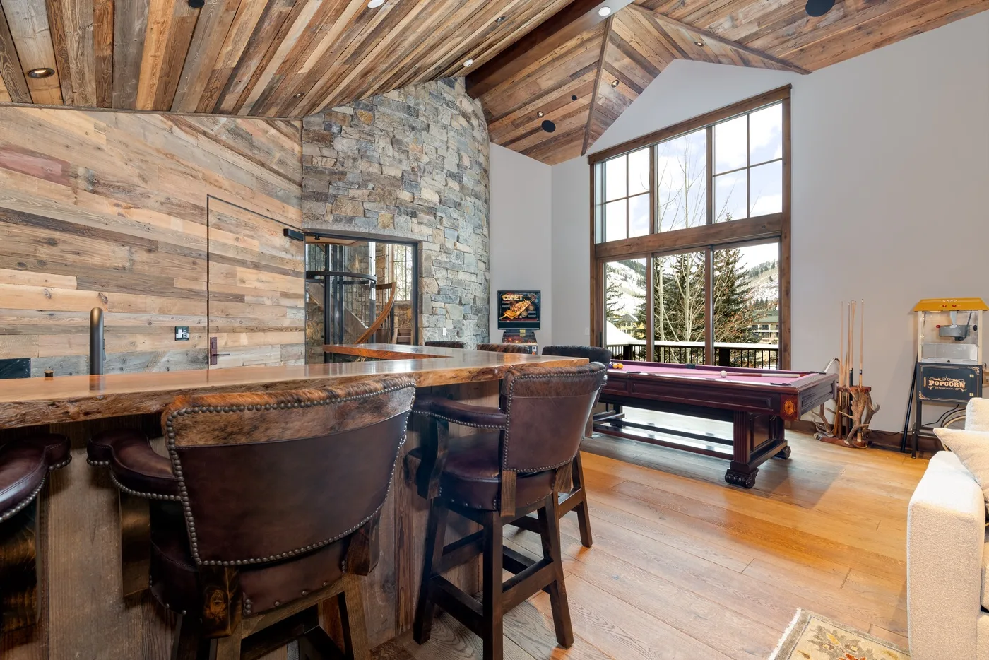 A rustic living room with a large bar and a pool table beside tall windows.