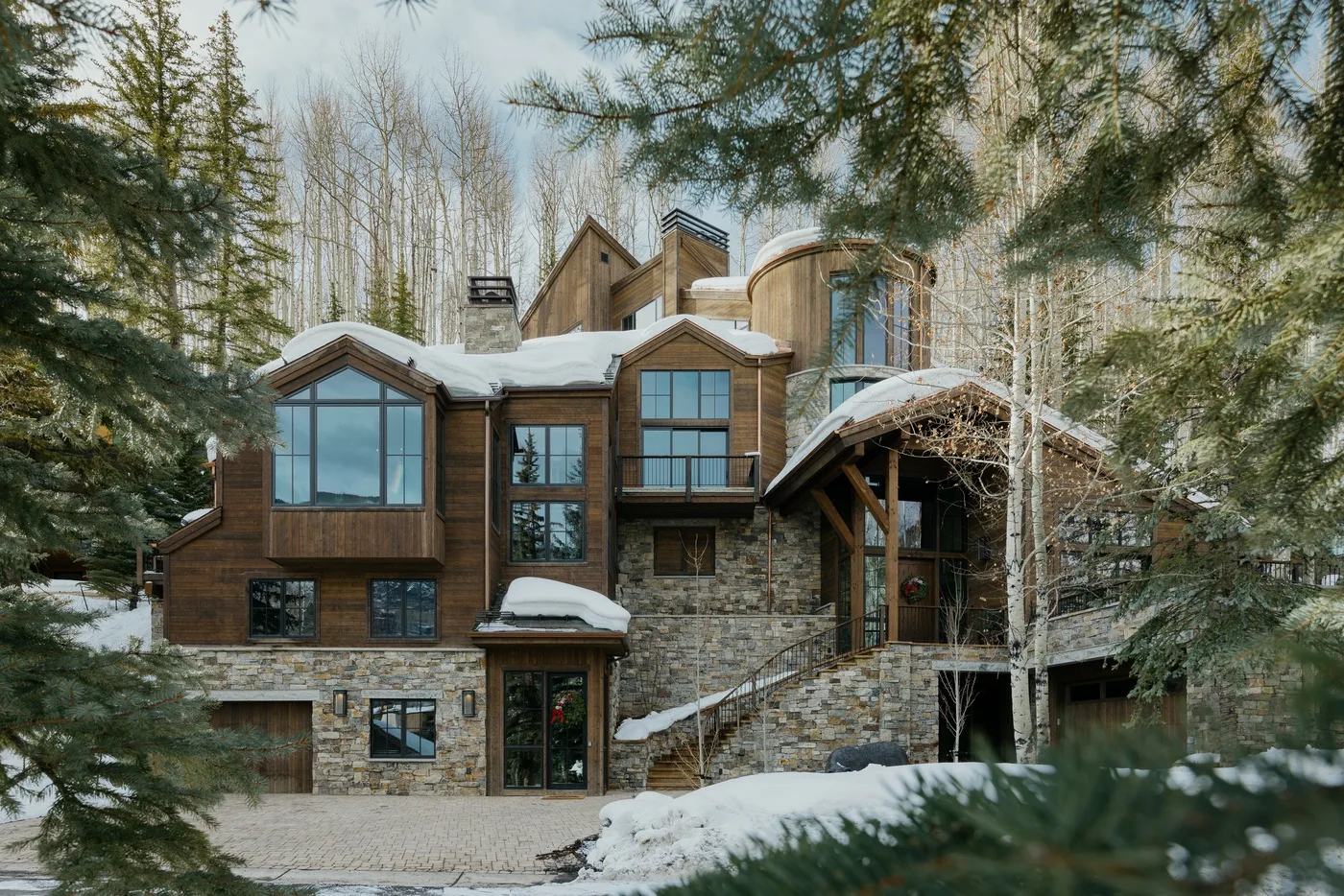 A large lodge-style house with stone walls and wood siding sits among snow-covered trees.
