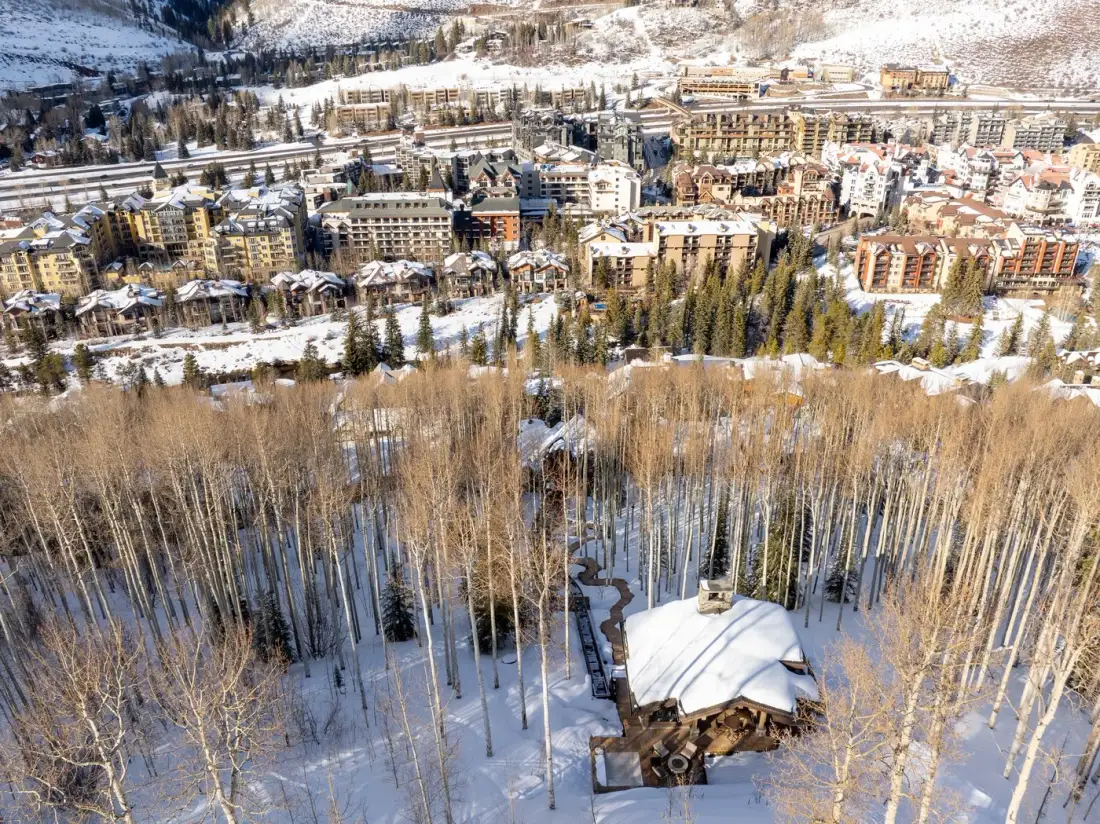 A snowy mountain town sits beyond a wooded slope, with a cabin in the foreground.