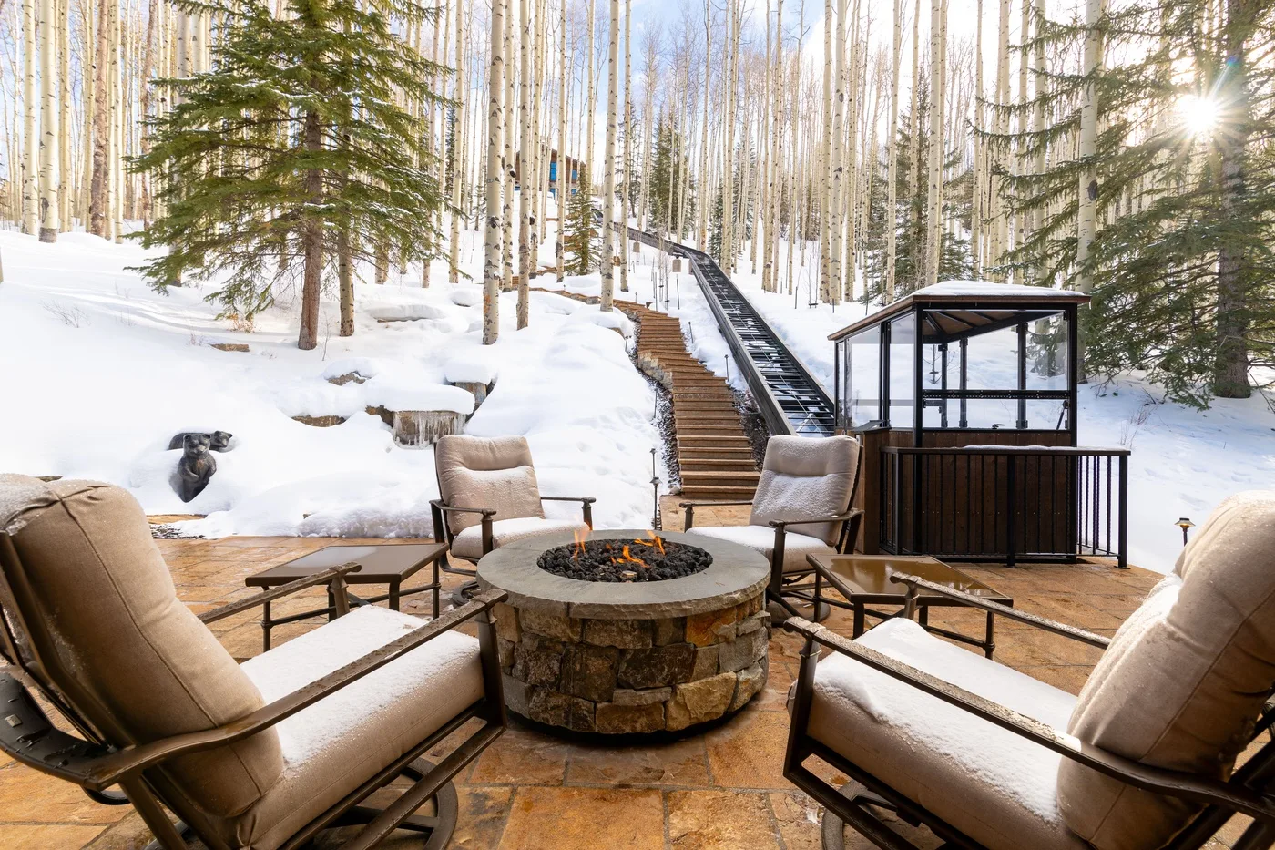 A snowy forest deck with outdoor seating, a fire pit, and stairs rising through the trees.