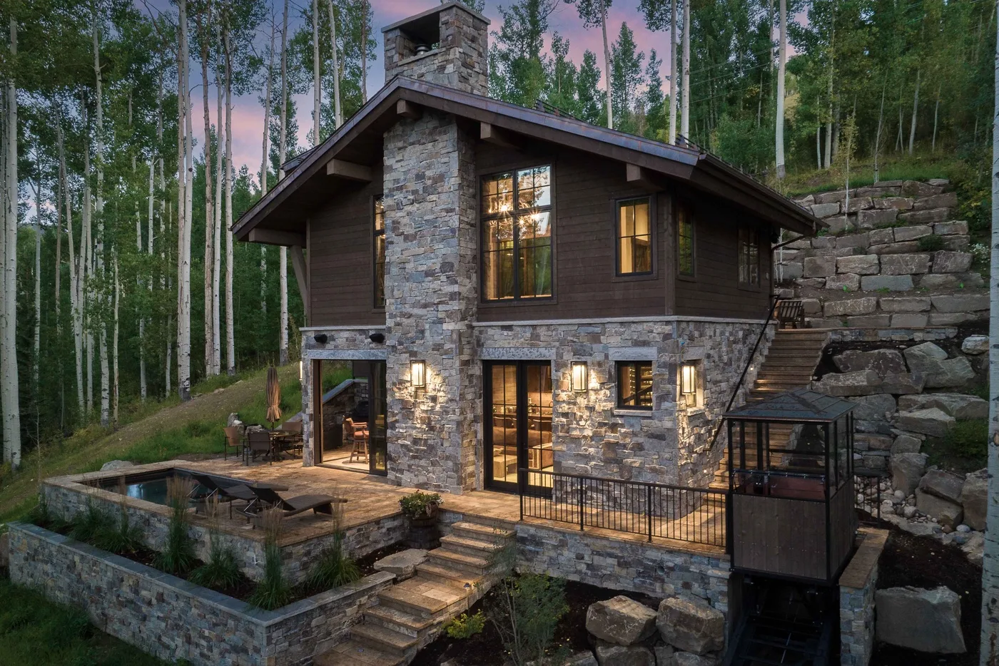 A stone-and-wood house sits among trees, with a lit patio and stone retaining wall.
