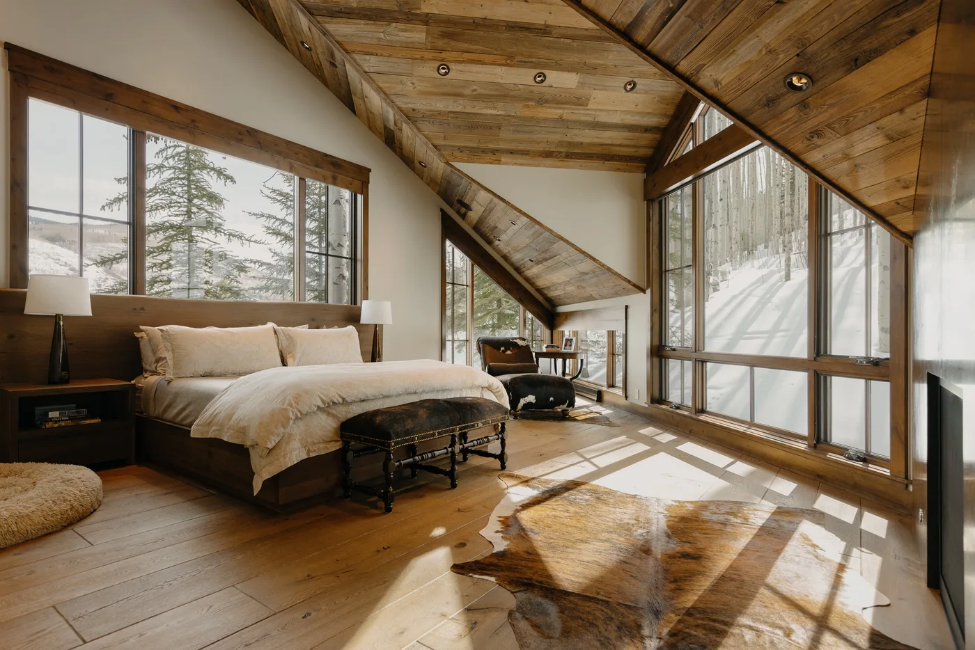 A bedroom with a bed beneath a sloped wooden ceiling and large windows.