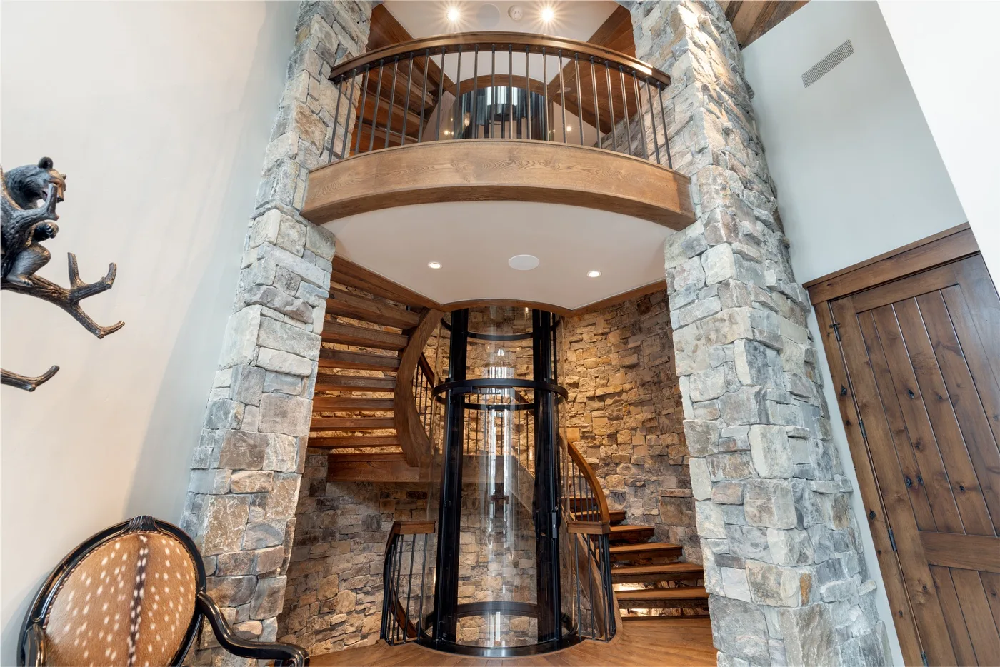 A stone interior features a curved staircase and a glass elevator beneath a balcony.
