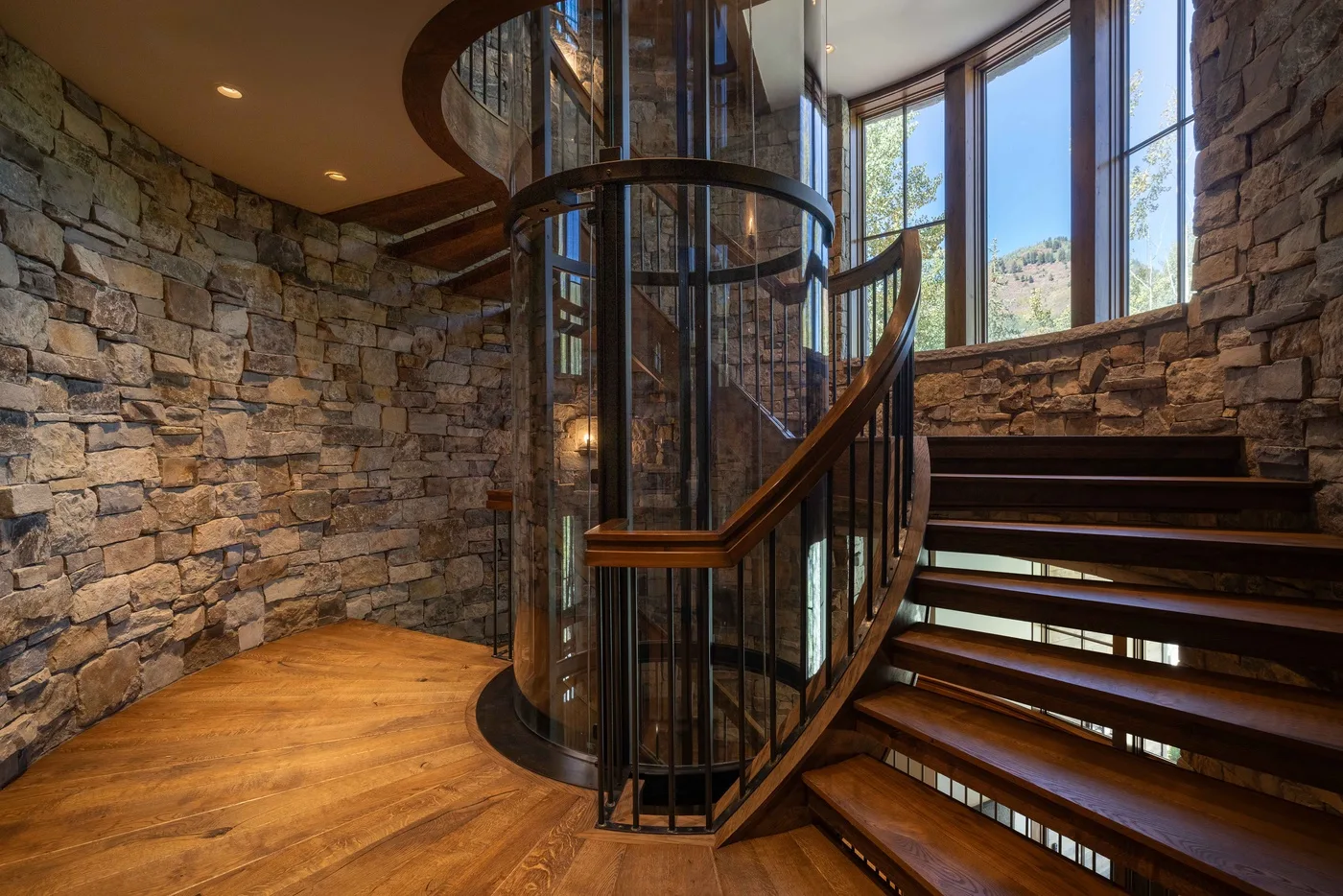 A curved wooden staircase wraps around a glass elevator in a stone-walled interior.