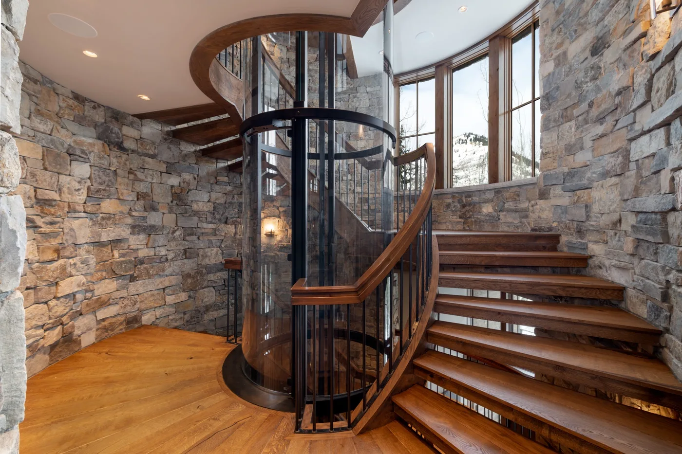 A curved wooden staircase rises beside floor-to-ceiling windows and stone walls.