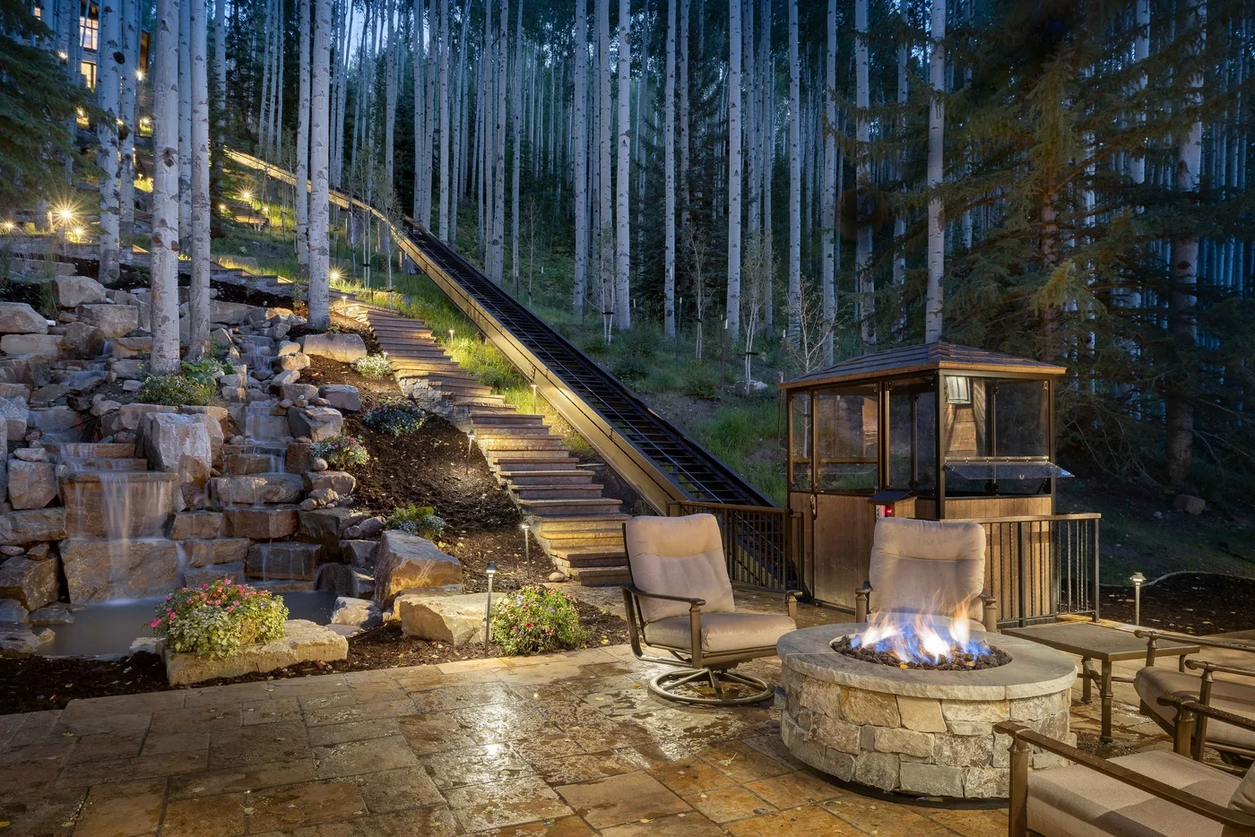 An outdoor patio with a stone fire pit beside a wooded hillside.
