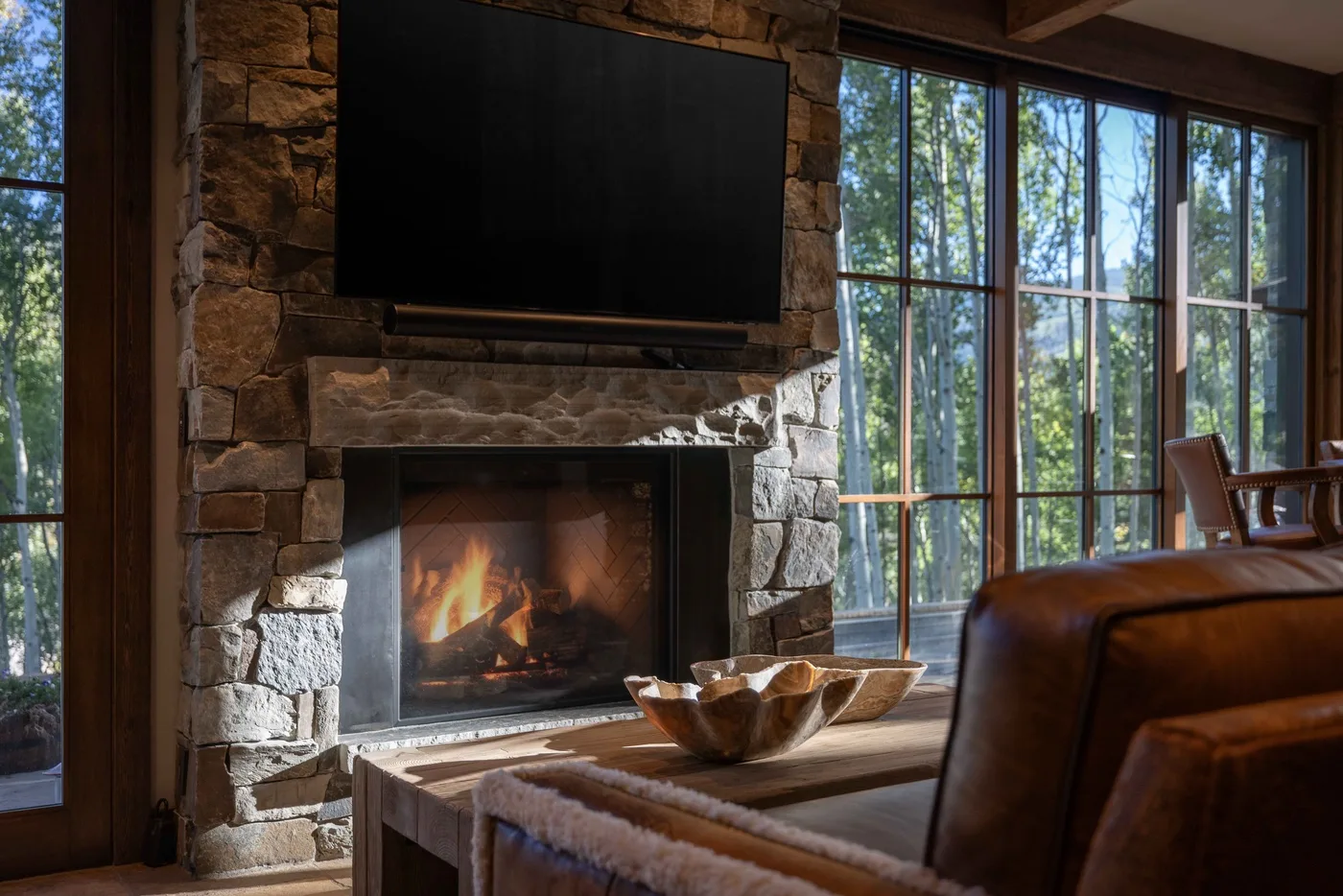 A living room with a stone fireplace and large windows overlooking trees.