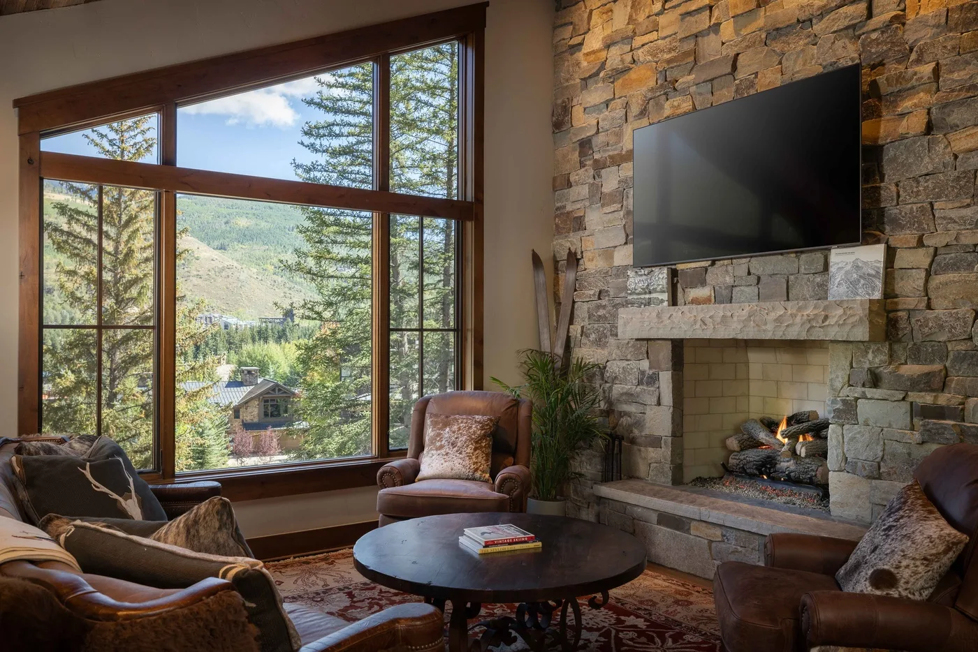 A living room with a stone fireplace, seating, and large windows overlooking trees.