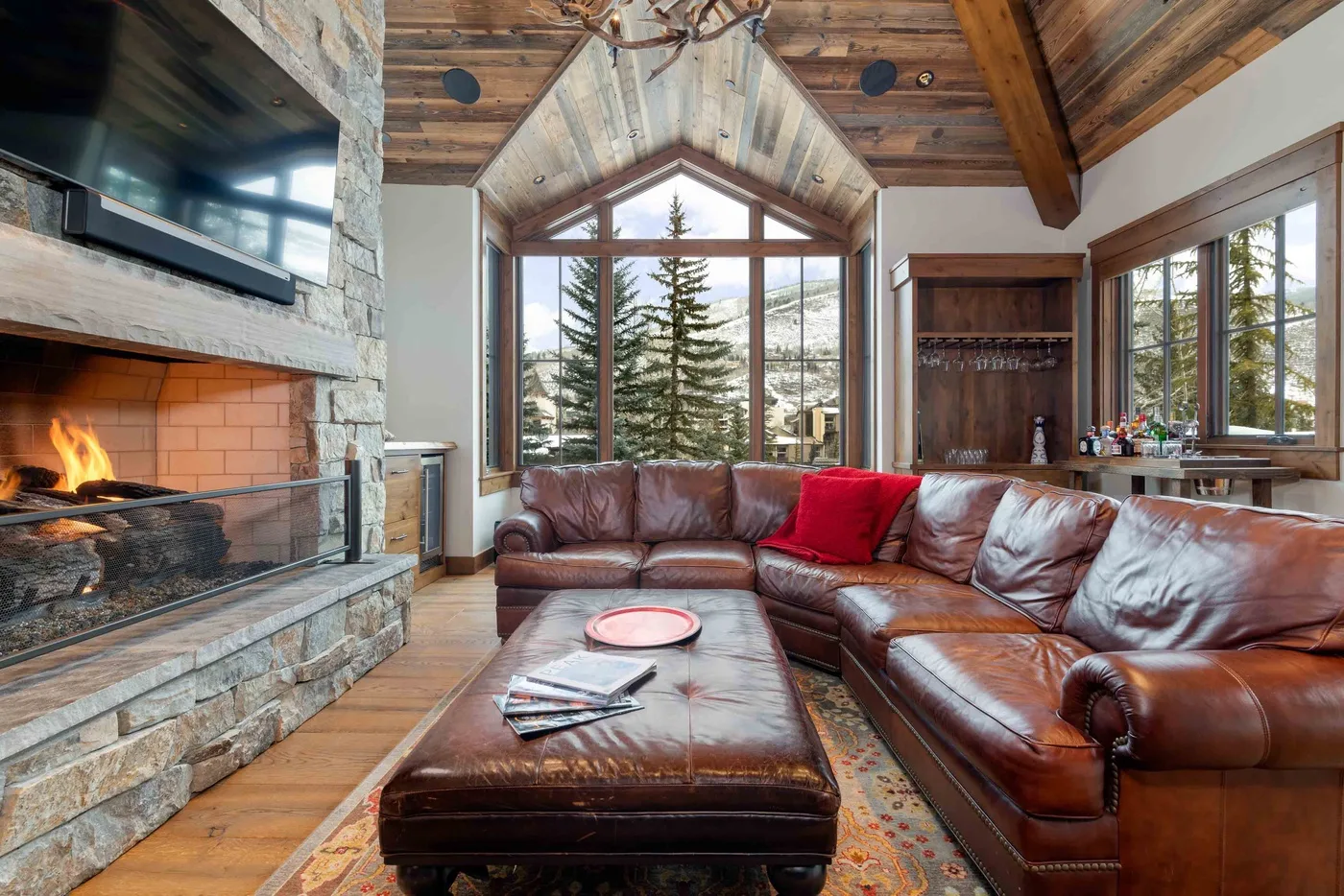 A rustic living room with leather seating, a stone fireplace, and tall windows.