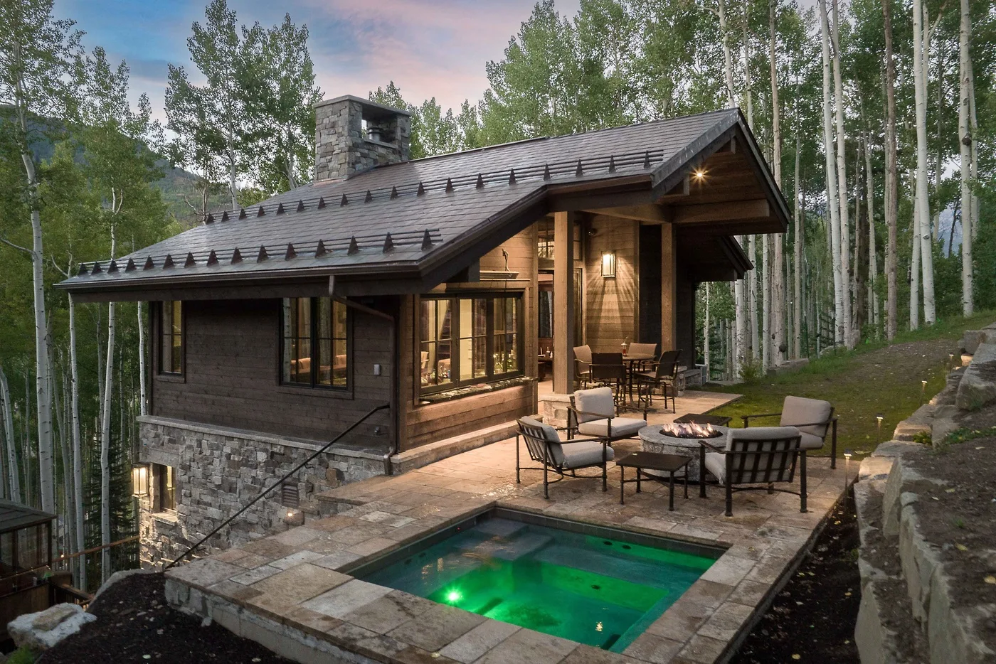 A woodland cabin with an outdoor patio, seating, and an illuminated pool.