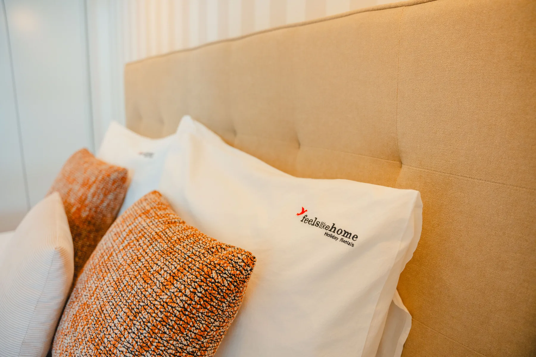Dormitorio con almohadas blancas y cojines estampados naranjas contra un cabecero acolchado.