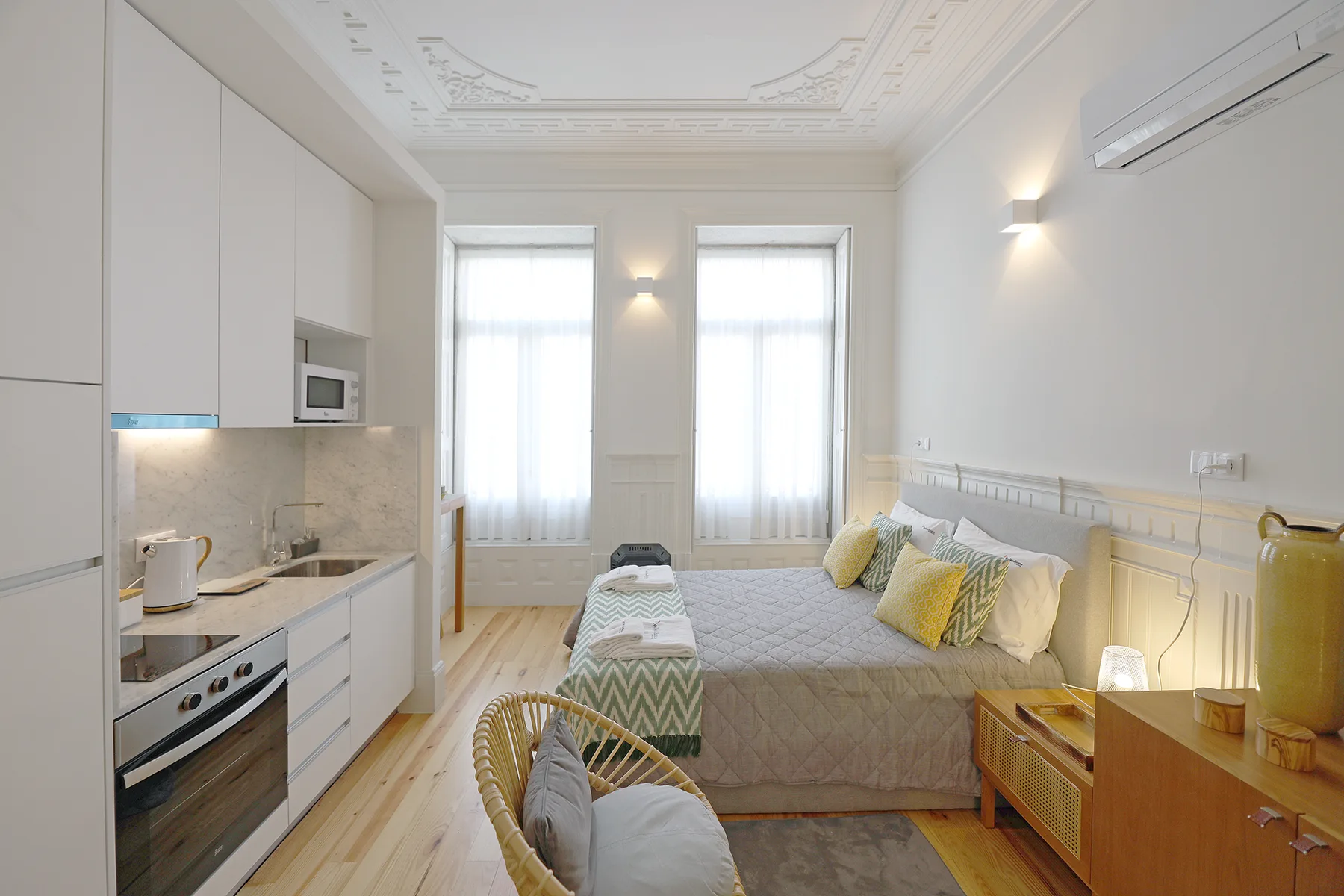 A studio room with a bed, kitchenette, and tall windows.