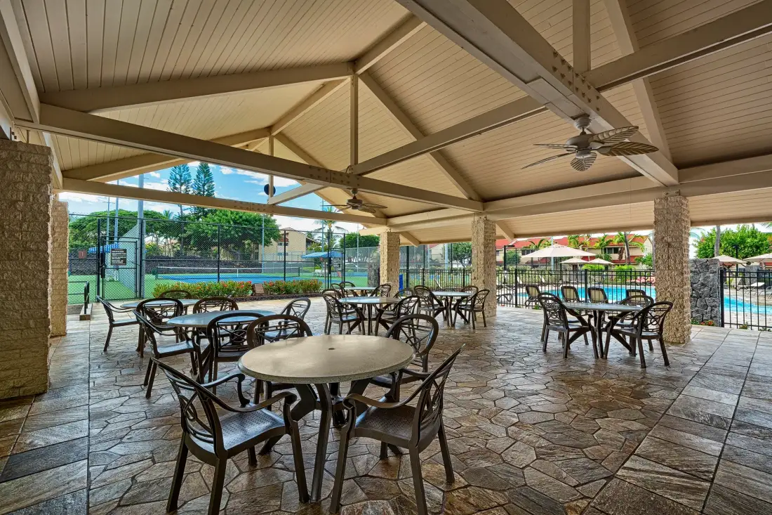 A covered outdoor dining area with round tables and chairs beside a pool.