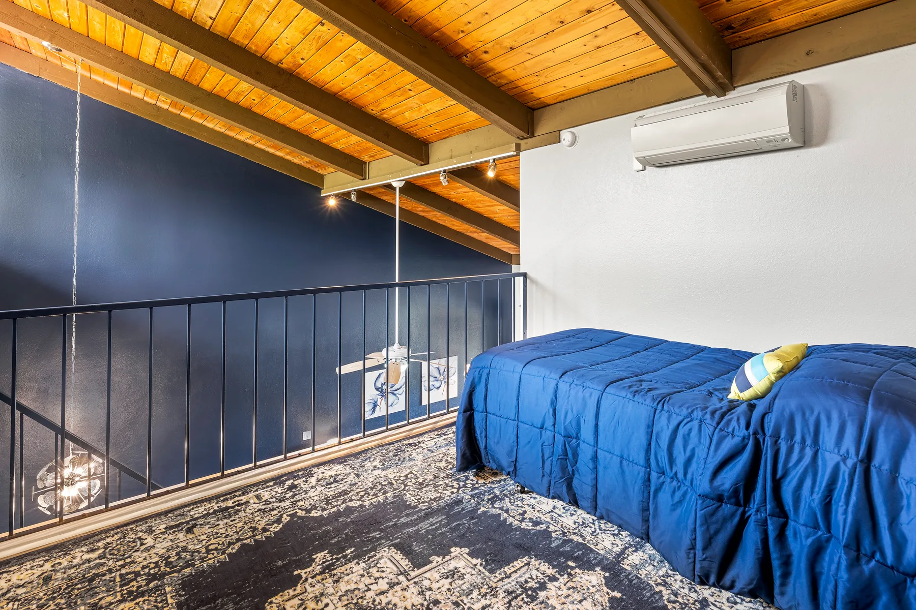 A bedroom with a blue bed beside a dark metal railing under a wood-paneled ceiling.