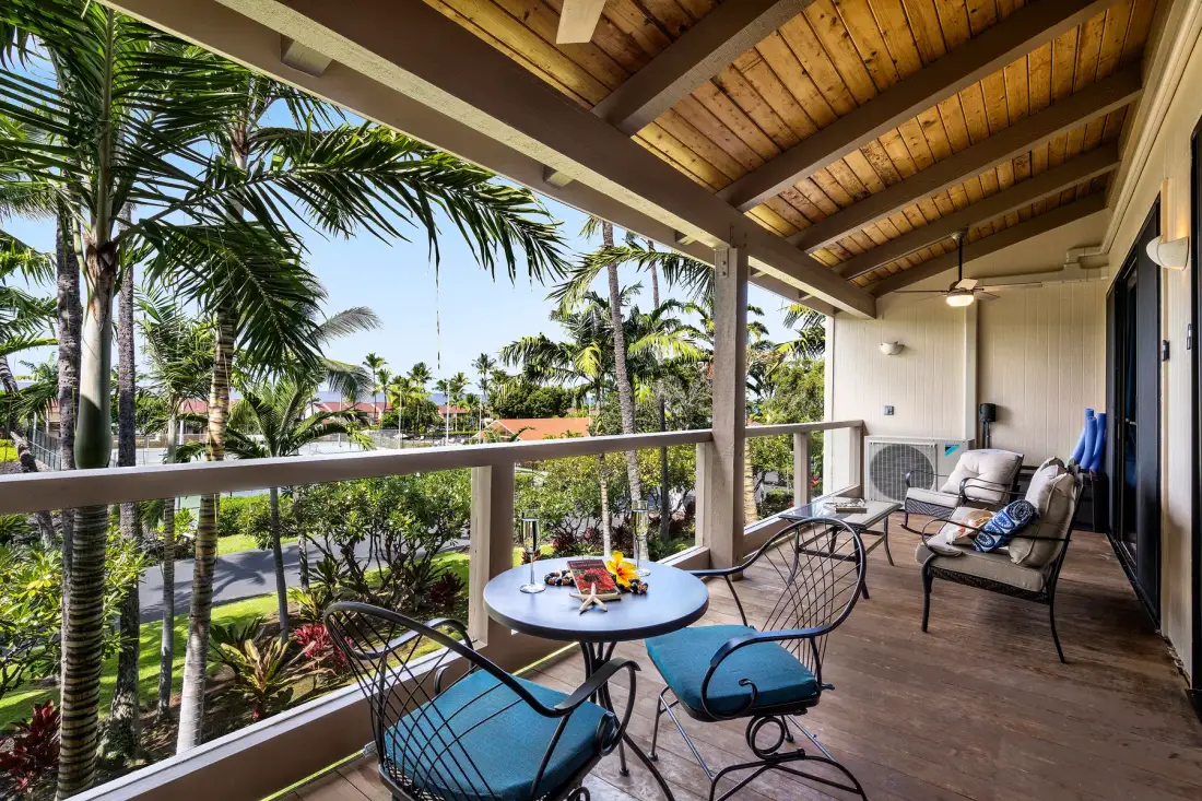 A covered balcony with seating and a small round table overlooks palm trees.