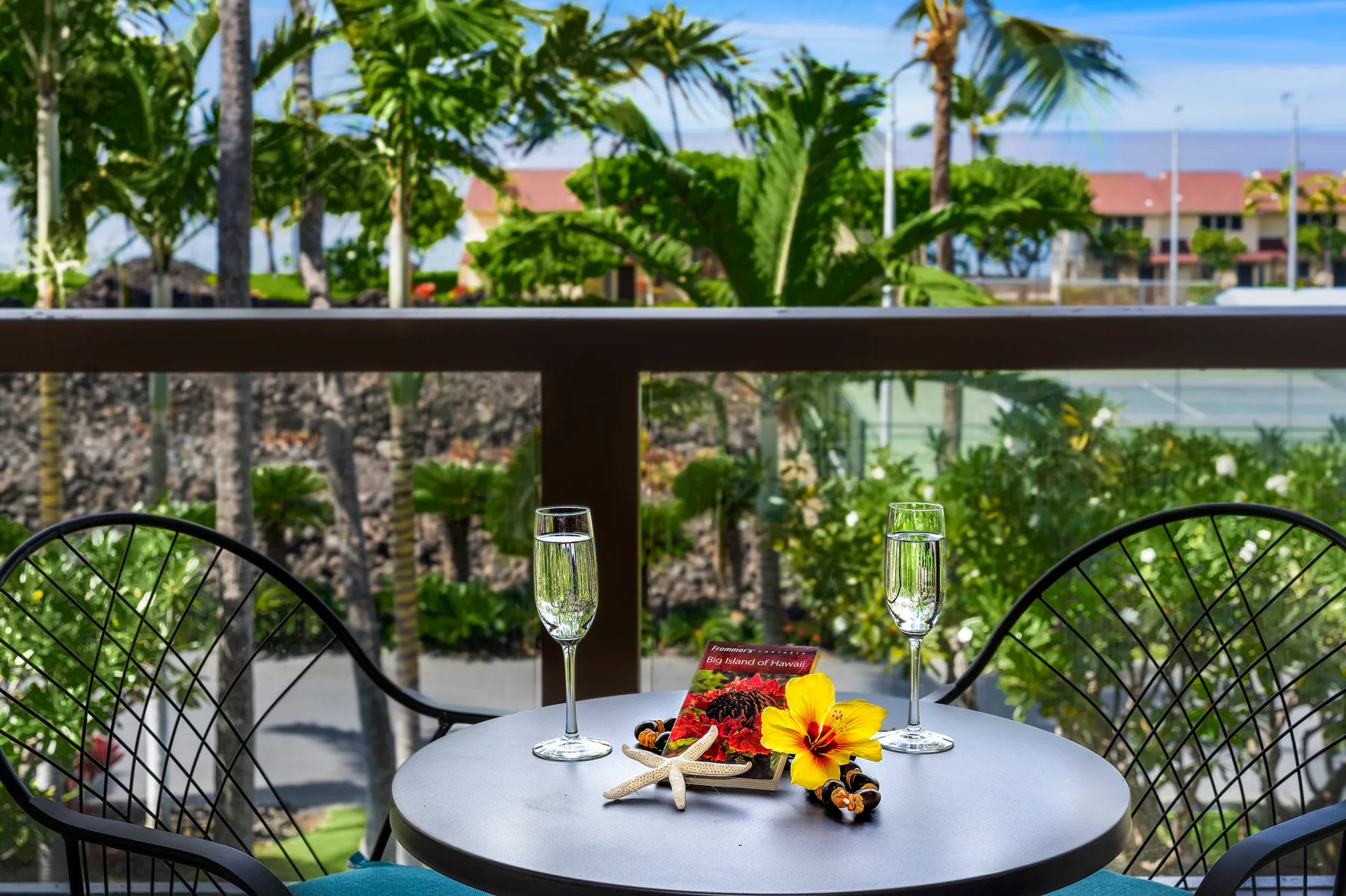 A balcony table with flowers and two glasses overlooks palm trees and greenery.