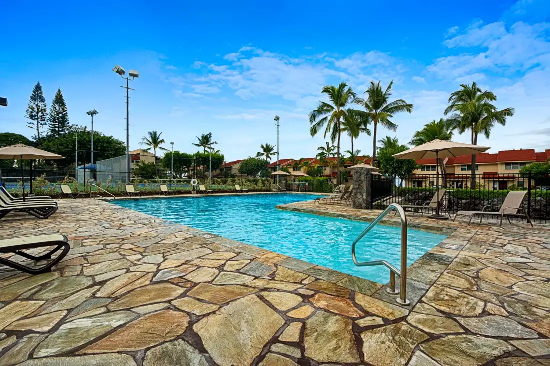 An outdoor swimming pool is surrounded by stone paving, lounge chairs, and palm trees.
