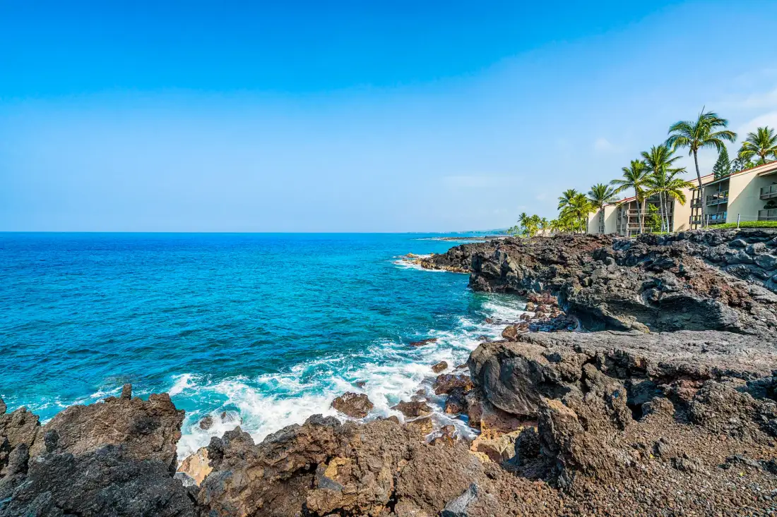 Rocky coastline beside turquoise ocean water and palm-lined buildings.