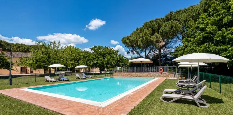 An outdoor swimming pool is surrounded by loungers, umbrellas, and green trees.