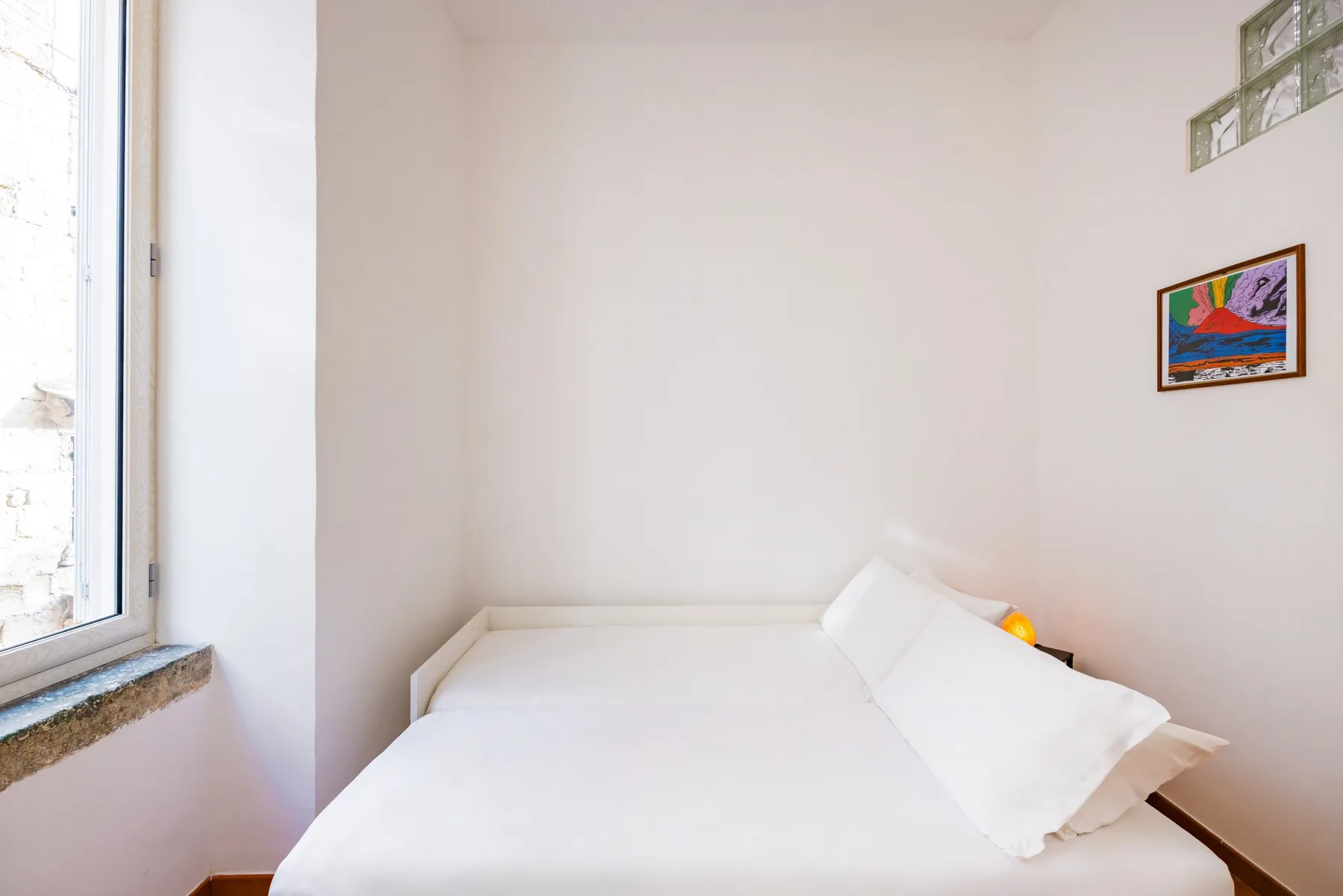 A bright bedroom with a white bed beneath a small framed picture.