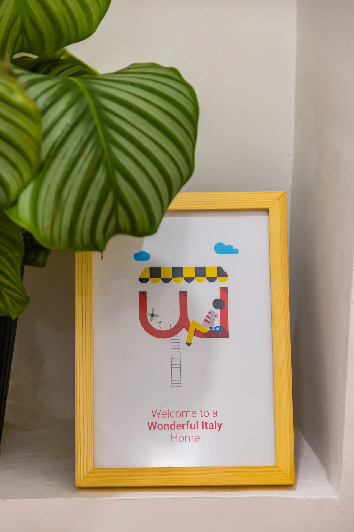 A framed colorful illustration leans against a wall beside a leafy plant.