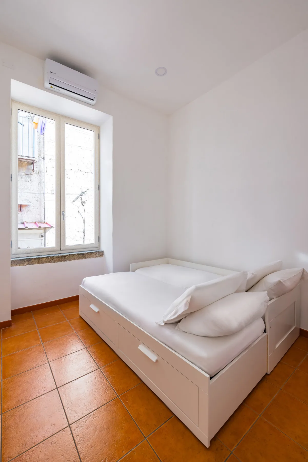 A bright bedroom with a white bed beside a tall window.