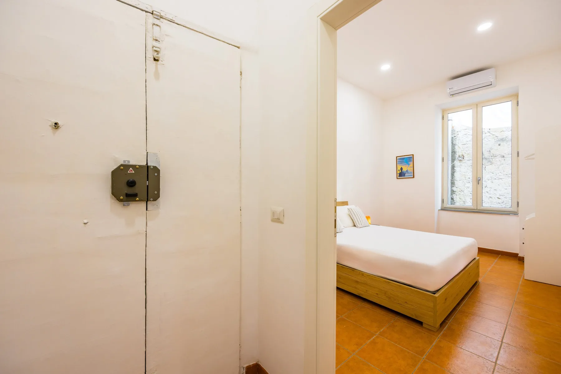 A bedroom with a bed, a window, and terracotta-colored floor tiles.