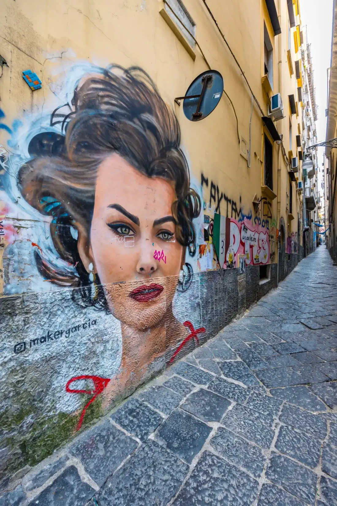A narrow stone-paved alley runs past a large portrait mural on the wall.