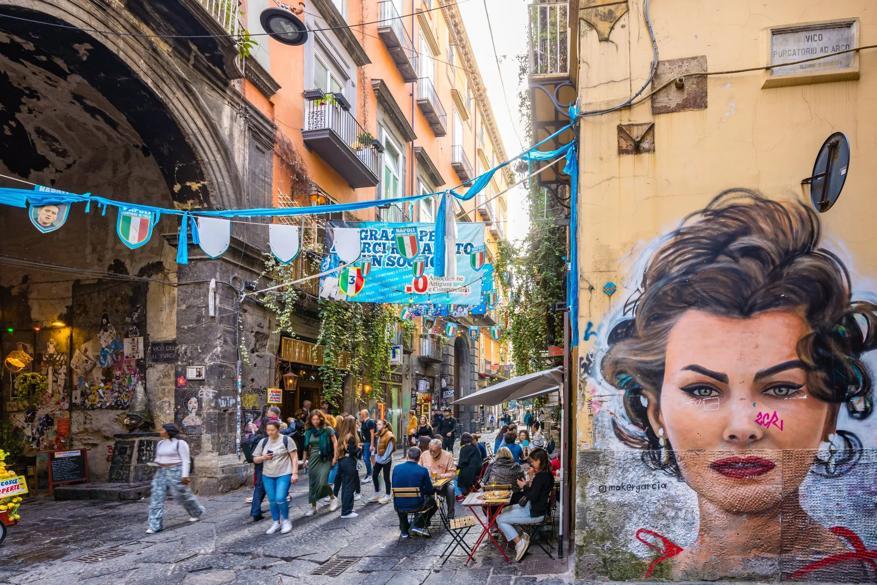 A narrow city street is lined with shops, pedestrians, and colorful buildings.