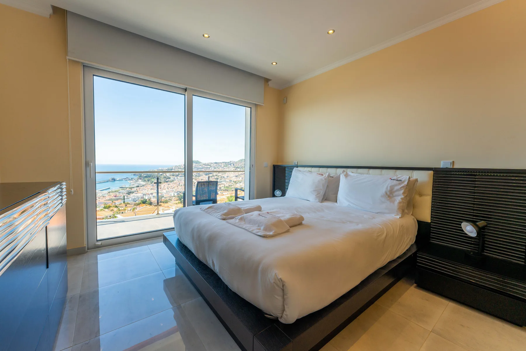 Un dormitorio con una cama bien arreglada junto a una gran ventana con vistas a la costa.