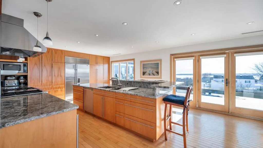 A bright kitchen with wooden cabinetry, stone counters, and windows overlooking the water.