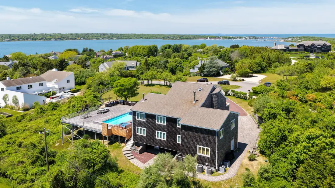 Aerial view of a dark house surrounded by greenery, with a pool and water in the distance.